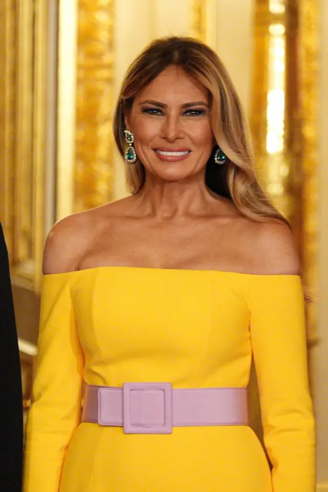 A close up vertical shot of a smiling Melania Trump in a yellow Carolina Herrera gown with a pink belt