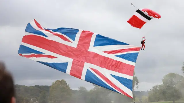 A paraglider with the Union Jack flag behind them descends towards a treeline