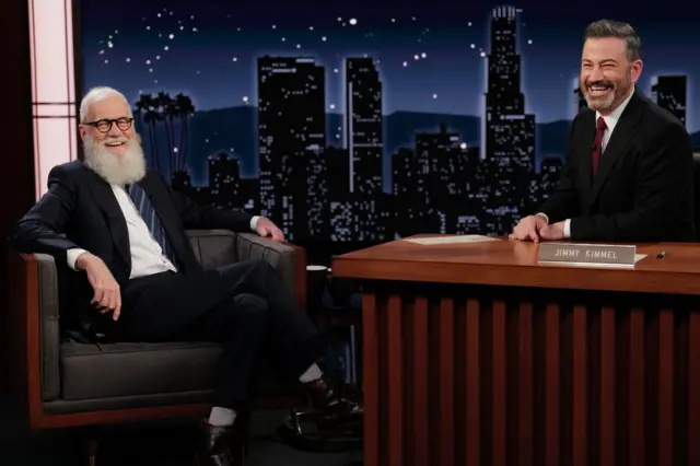David Letterman, left, seated with his leg crossed, laughs with Jimmy Kimmel who is seated behind a brown desk.