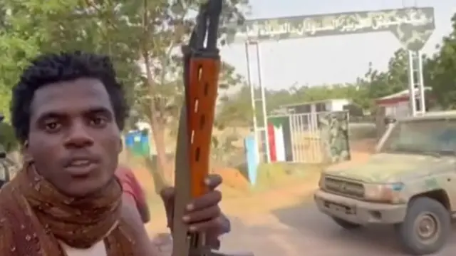 A RSF fighter holding a machine gun and wearing a scarf, there is a truck and a some gates with the Sudanese flag on it in the background