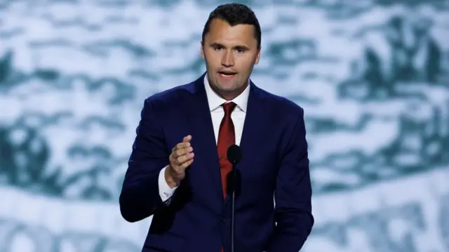 Charlie Kirk, is a dark blue blazer with red tie, speaks into a microphone placed on a lectern in front of a light blue backdrop.