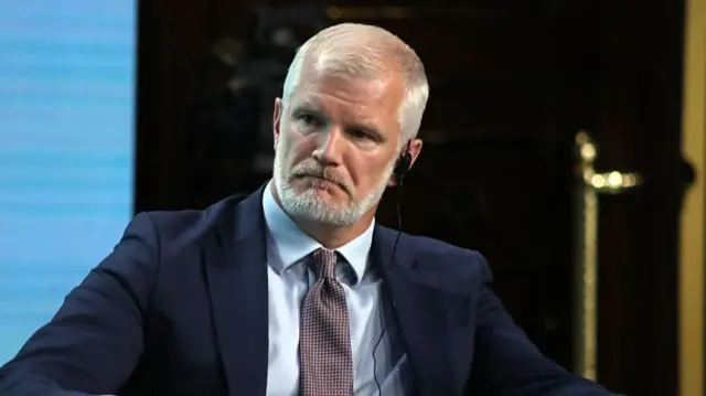 A man in a white shirt and navy-blue suit jacket looks to the left of the camera. He has white hair and a beard.