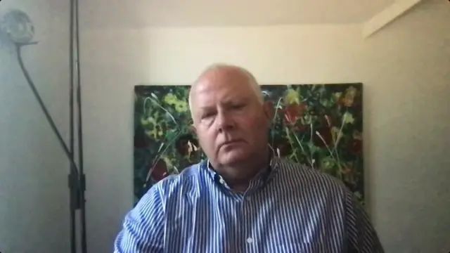 A man staring down the lens, sitting in a room. He is wearing a white and blue striped button shirt. A lamp sits to his right and a colourful picture is hung up behind him.