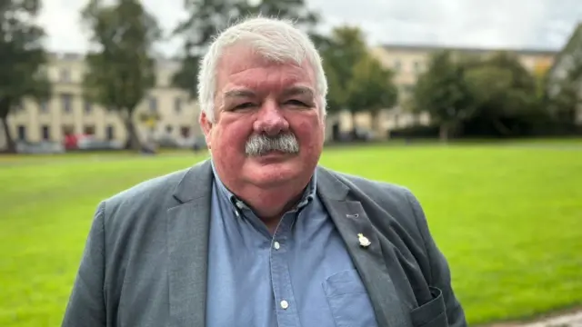 A man with white hair, a white moustache, a blue shirt and grey jacket. On his lapel is a pin.