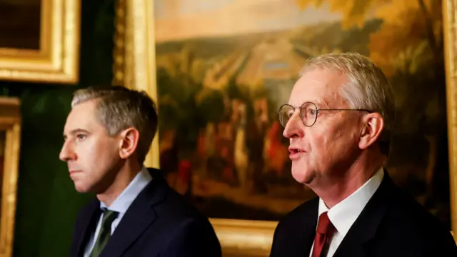 Harris and Benn speaking at the podium. Both are in front of grand paintings.