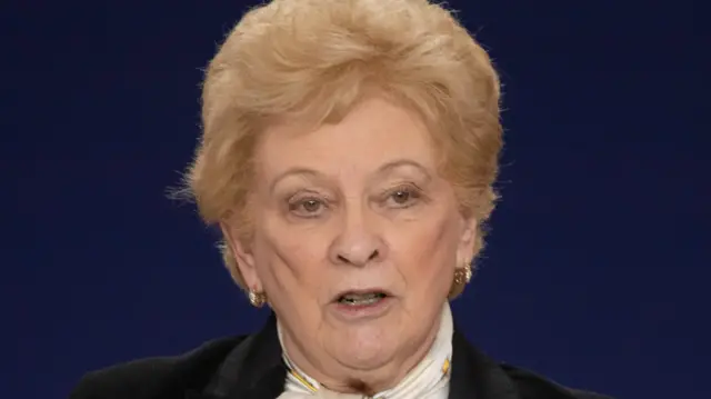 Nuala O’Loan is wearing a black jacket with a yellow scarf. She has short blonde hair.