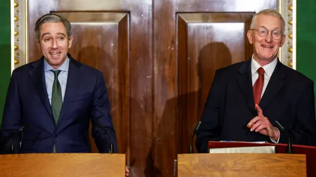 Harris and Benn smiling at the podiums infront of a wooden door. Harris has a green tie on and Benn has a red tie on.