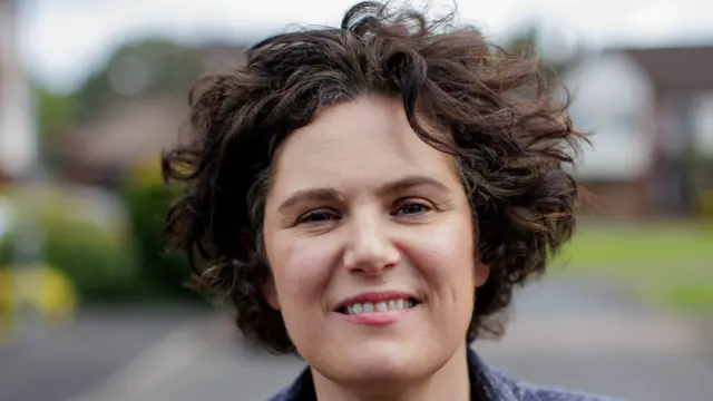 Claire Hnnna has curly short brown hair. She is smiling at the camera.