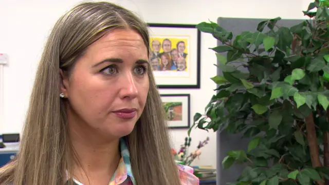 a woman looks to the left of frame, she is stood beside a fern