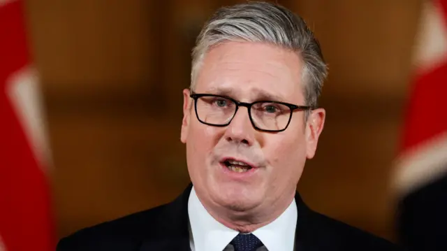 A close up shot of Keir Starmer wearing a black suit and white collar, with two union jack flags blurred in the background behind him