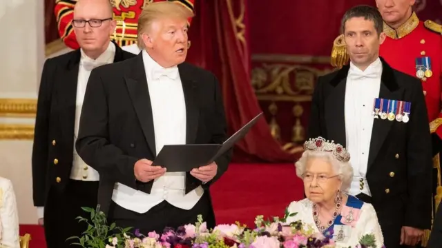 Trump is standing up holding a folder and speaking while the Queen is sitting down and listening