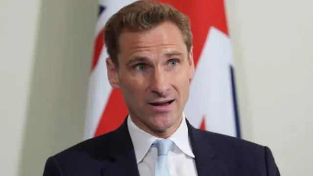 Chris Philp has short, dark blonde hair and is standing in front of a Union Jack flag, looking off to the side.