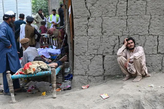 An Afghan man squats in front of a wall. Around the corner are beds with people sitting or lying on them.
