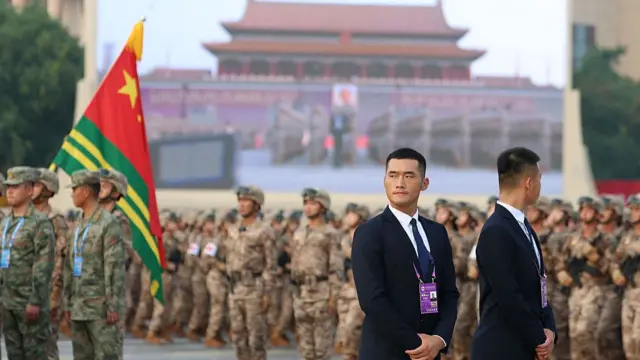Two men in suits look in different directions. Behind them are soldiers standing in neat rows.
