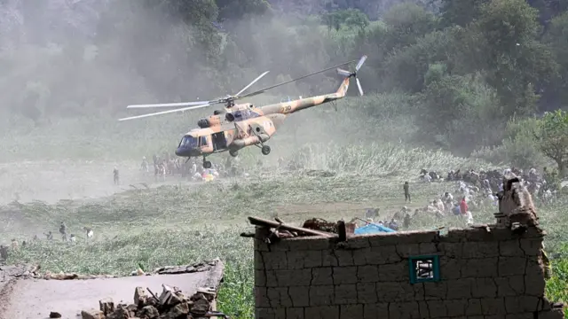 A helicopter flies above damaged houses and an open green field
