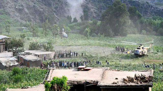 A wide shot of a helicopter landing onto a green field, as people crowd around.