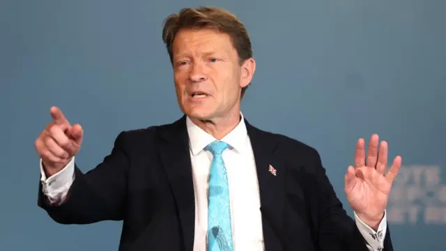 Tice, in suit and blue tie, points