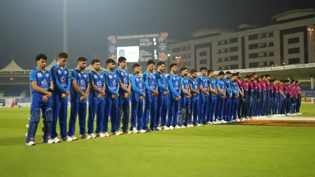 Players stand in a row on the cricket field, with lowered heads and crossed hands.