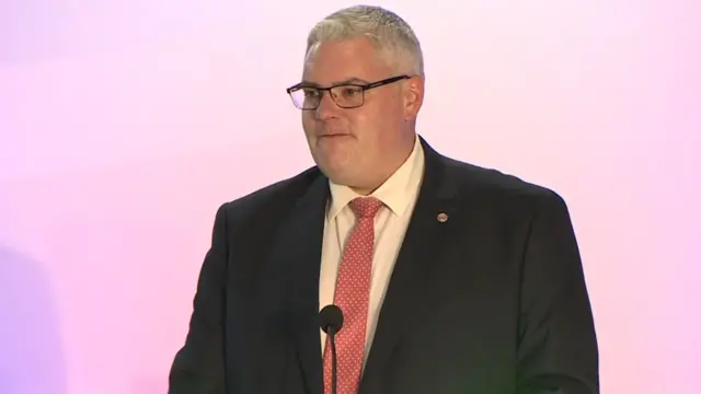 A man wearing a suit and glasses stands at a microphone,. He has white hair, and the background behind him is pink.