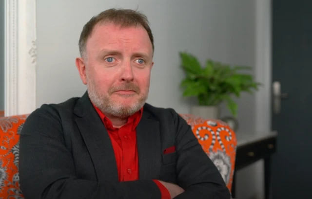Chris McCausland wearing a red shirt with his arms crossed