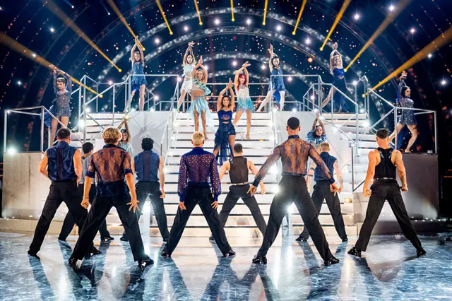 The back of two lines of male professional dancers, who are facing nine professional female dancers coming down white steps on the Strictly stage