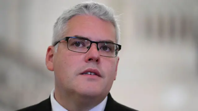 A man with white hair and glasses. He appears to be wearing a suit.