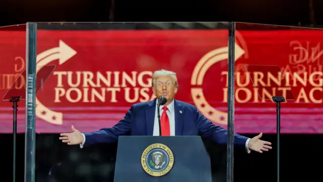 Trump stands behind a glass screen as he speaks at Charlie Kirk's memorial. A sign that reads "turning point USA" appears behind him.