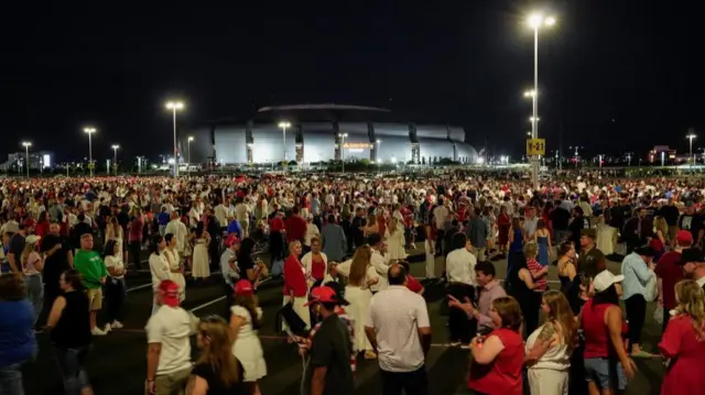 A crowd of people stand waiting outside the stadium. The sky is dark and a flood light is on