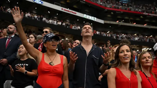 Um homem fecha os olhos e mantém as palmas das mãos abertas, aparentemente rezando, enquanto a multidão no estádio pode ser vista atrás dele