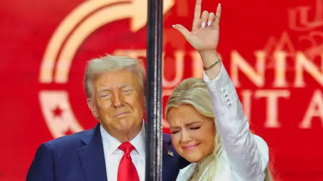 A man with white hair has his arm around a blonde woman in a white suit. They both have their eyes closed, and the woman holds her hand in the air in a religious hand gesture.