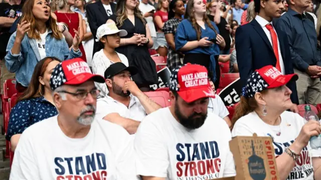 Três pessoas sentam-se nas arquibancadas do estádio usando chapéus com os dizeres "Make America Great Again" - outras pessoas ficam atrás delas