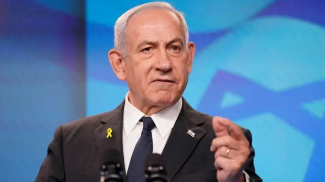 Netanyahu speaking into two microphones at an event and pointing with one hand.