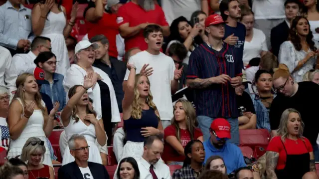 Pessoas nas arquibancadas do estádio - algumas estão de pé, cantando e agitando os braços