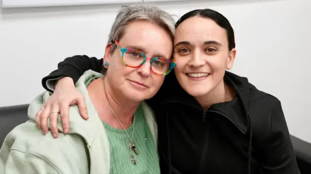 Mandy Damari, who has short grey hair and wears a sage hoodie and green top, is embraced by her daughter Emily, whose black hair is tied back and wears a black hoodie