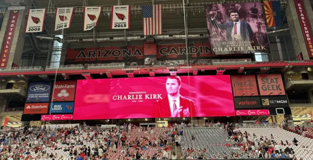 As pessoas começam a sentar-se nas arquibancadas do estádio, abaixo de uma faixa vermelha com os dizeres "Em memória de Charlie Kirk".