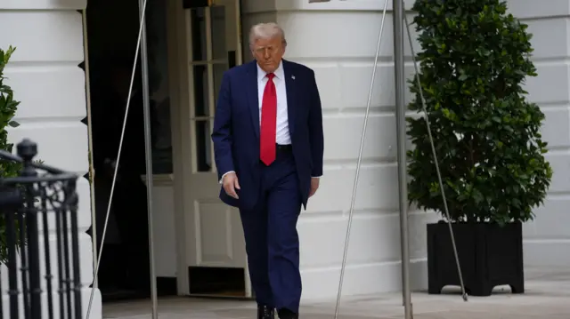 Trump dressed in a red tie and a navy blue suit walks towards the camera with his head bowed down