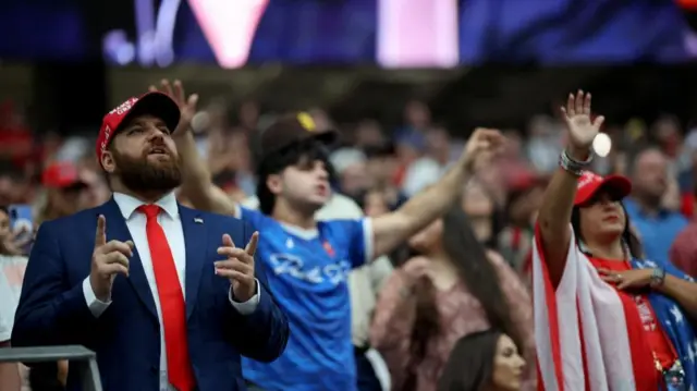 Um homem usando um boné da Maga canta na frente da multidão. Outros que estão perto dele também cantam e agitam os braços.