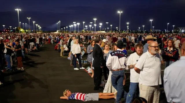A boy wearing a shirt with a U.S. flag pattern lies on the ground as people arrive, ahead of the morning sunlight, to attend the memorial service