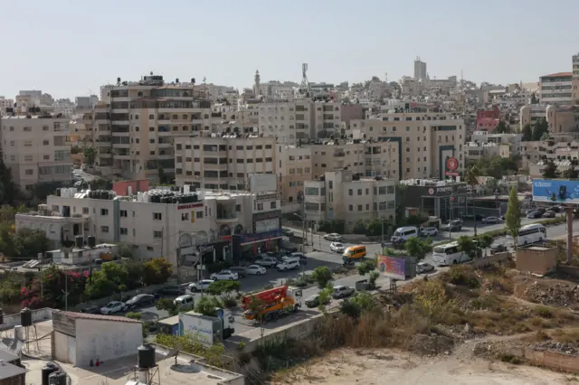 A wideshot of Ramallah, the Palestinian city. Sand coloured buildings and towers, and an earthy field.