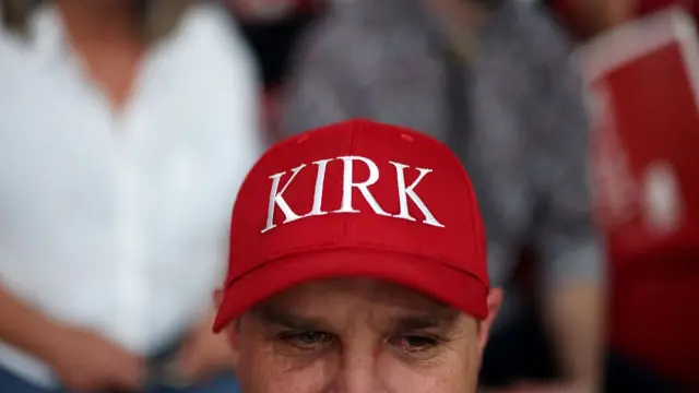 Um participante usando um boné vermelho com a inscrição "Kirk" sentado nas arquibancadas antes do serviço memorial para o ativista político Charlie Kirk no State Farm Stadium