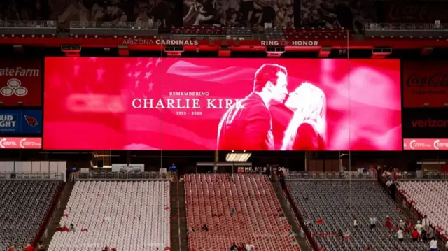 A large red screen reading "Remembering Charlie Kirk 1993- 2025" with an image of Charlie and Erika Kirk. Some people have begun sitting in the stadium seats
