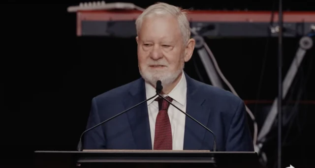 Larry Ann speaks from behind a podium. He is wearing a blue suit over a white shirt and red tie.