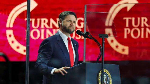 JD Vance stands at a podium against a red backdrop reading "Turning Point USA"