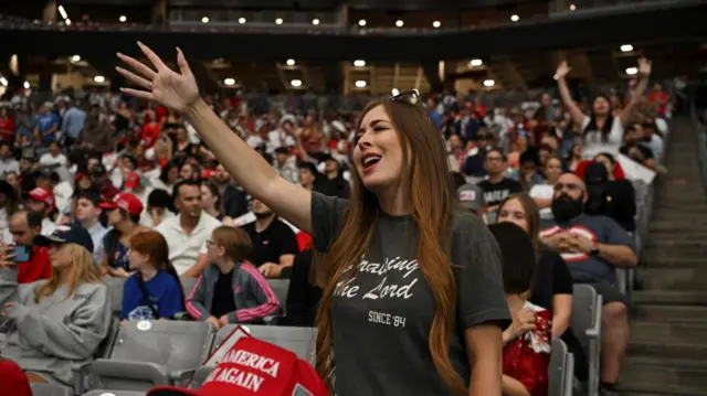 Uma mulher vestindo uma camiseta "louvando ao Senhor desde 1984" estende o braço, com os olhos fechados, com a multidão no estádio atrás dela