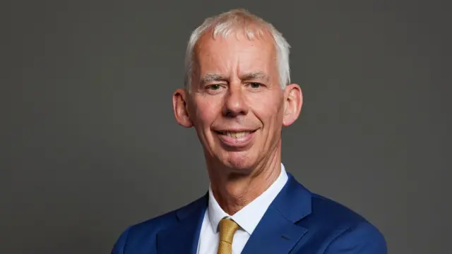 This is a close up photo of John Milne showing his head and shoulders. He is smiling. He has short white hair. He wears a navy blue suit, a white shirt and a gold tie.