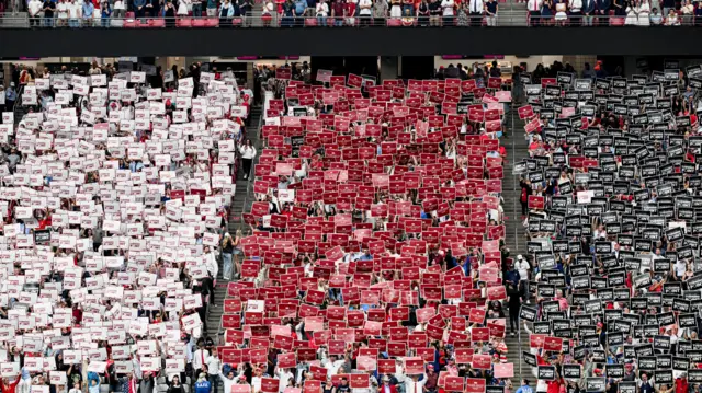 Three columns of supporters inside a stadium, holding up white, red, and black signs