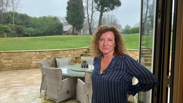 Sally Pavey, wearing a blue and white blouse, stands in her garden, with a set of garden furniture behind her.