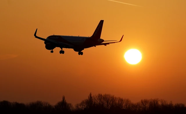A sunset image of a plane. The plane is a silhouette against an orange sky and bright yellow sun.