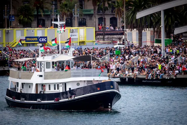 A boat with two levels sets sail from a dock with dozens of people waving them off, some waving Palestinian flags
