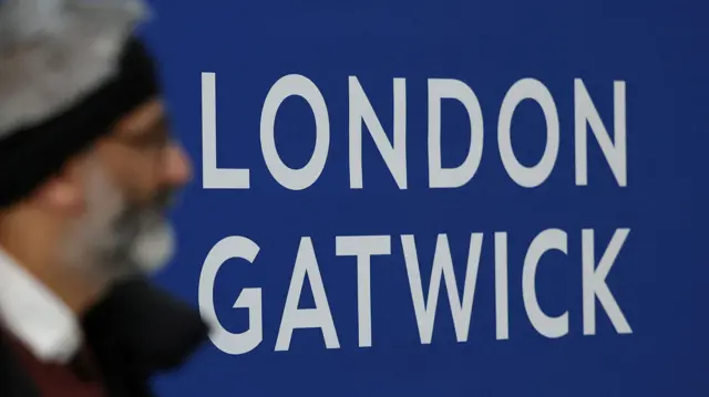 A passenger arriving at Gatwick Airport, with the sign for the airport in the background.